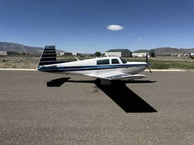 A white and blue Mooney aircraft, registration N231DD, parked on an asphalt ramp with hangars and mountains in the background.