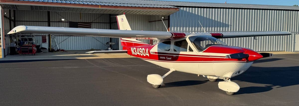 A Cessna Cardinal aircraft with red and white paint scheme is parked in front of a hangar.