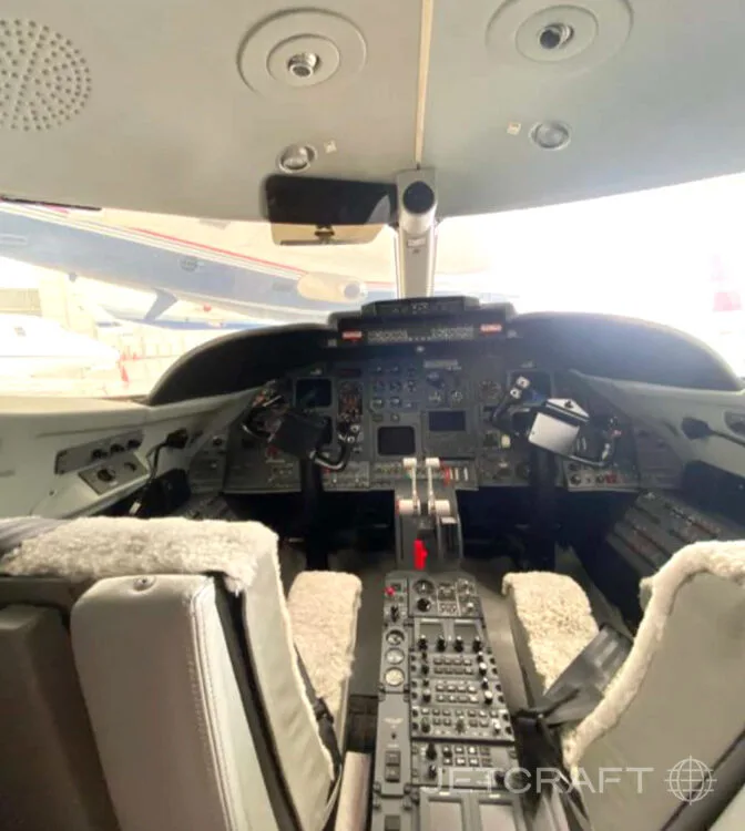 The cockpit of a business jet, showing the instrument panel, throttles, and two pilot seats with sheepskin covers.