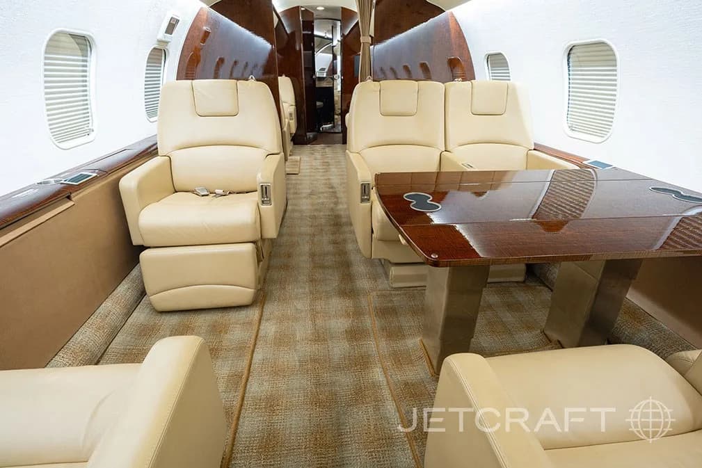 The aircraft cabin interior showing beige leather seats and a polished wooden table with cup holders.