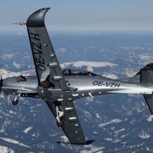 A silver CEAD aircraft flies over a snowy mountain range.
