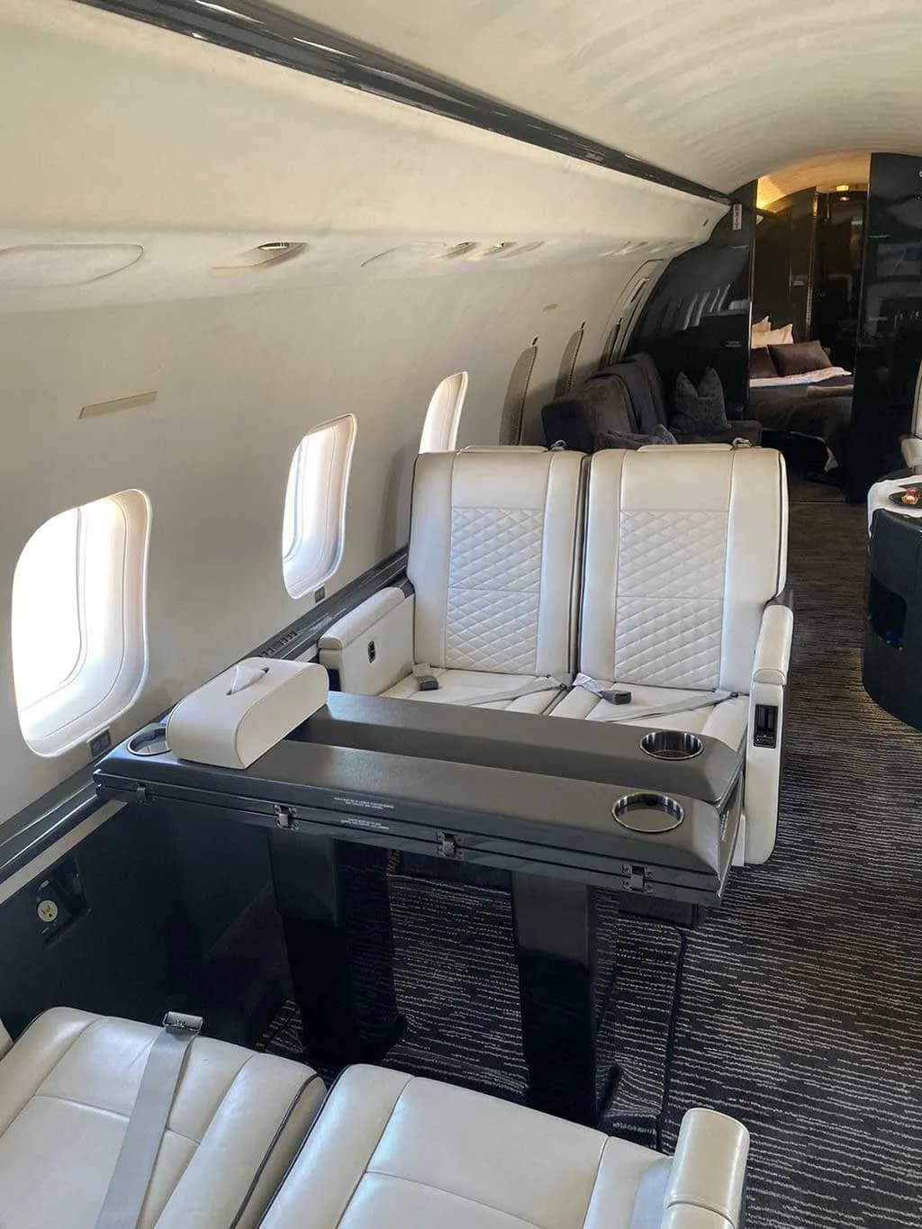 A view inside a private jet cabin, showing two white quilted seats facing a dark table with cup holders and a tissue box.