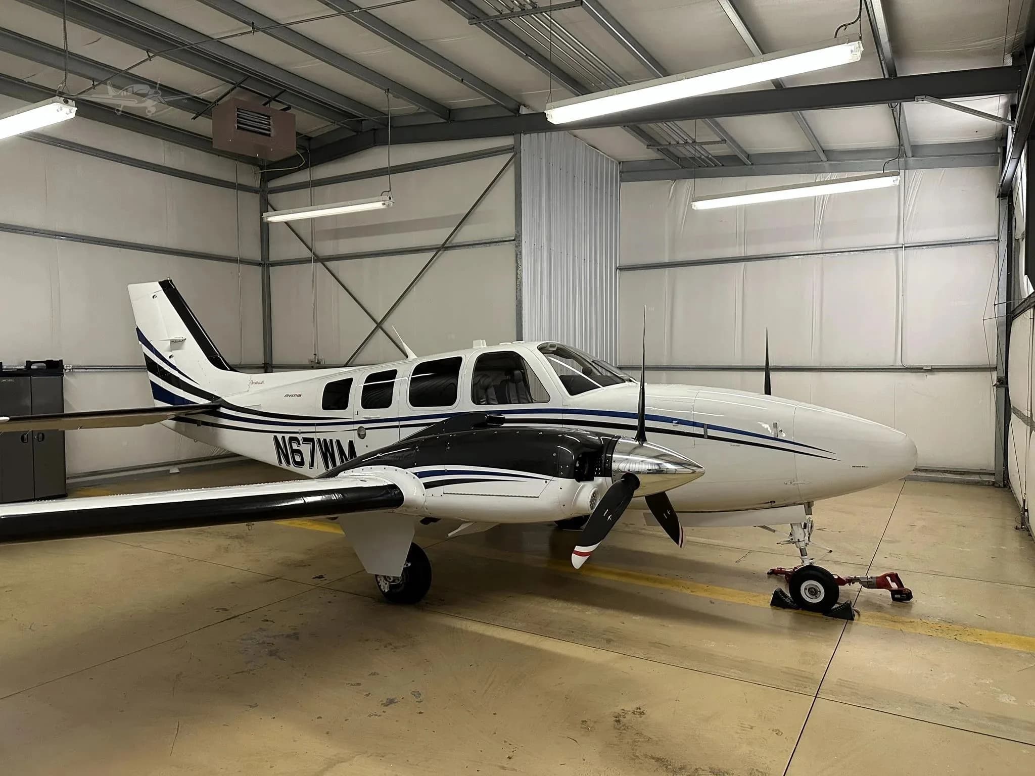 A white and blue Beechcraft Baron N67WM parked inside a hangar, viewed from the front-right.