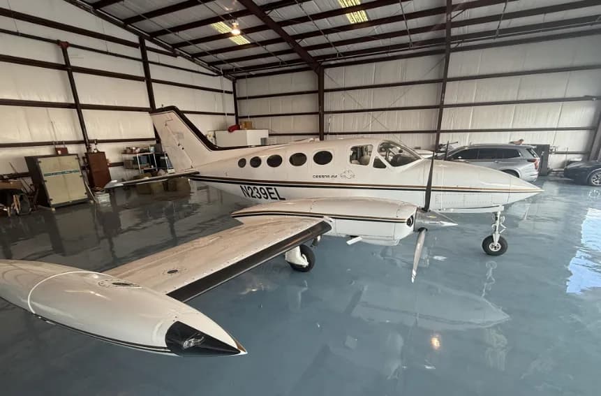 A white Cessna 414 aircraft with gold and black stripes and registration N239EL, parked in a hangar, viewed from the front-right.