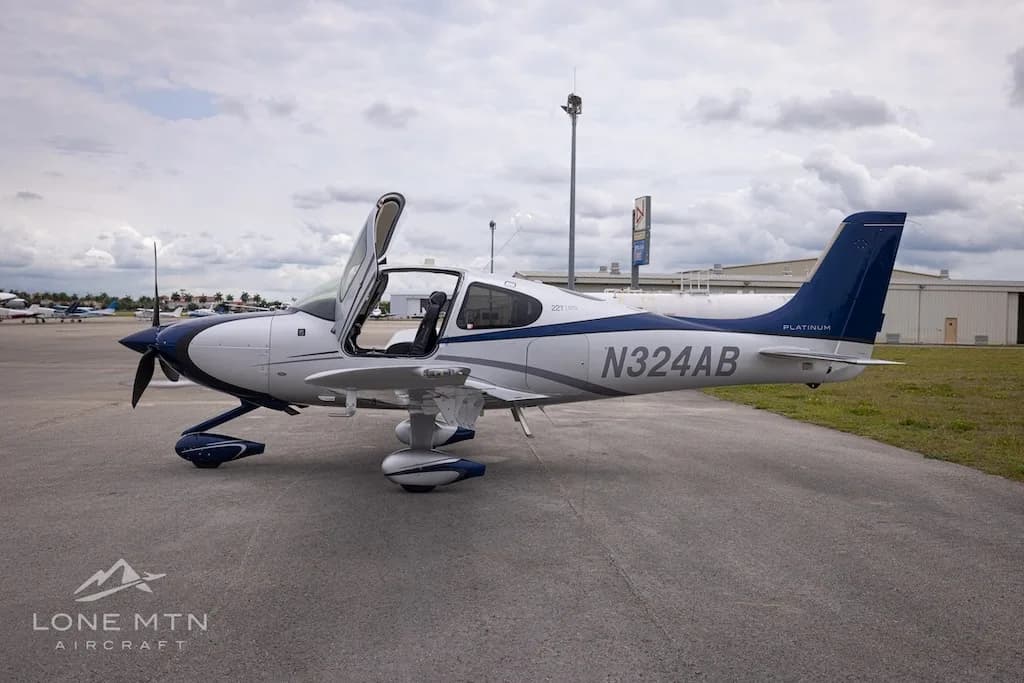 A white and blue Cirrus SR22T aircraft with open doors parked on an airfield, viewed from the side.