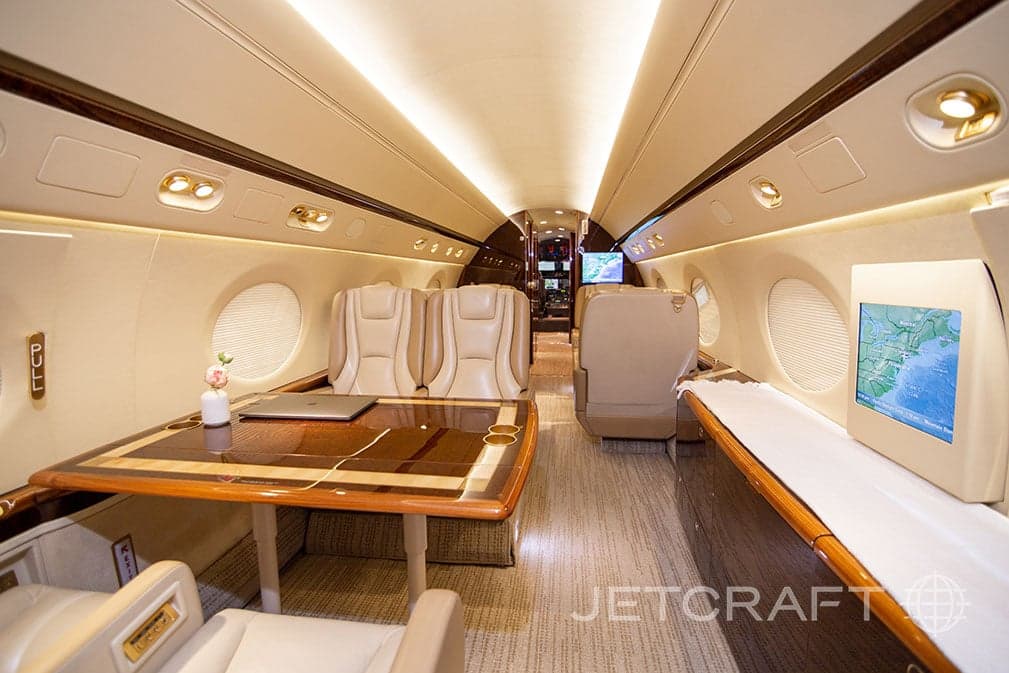 The spacious cabin interior of a Gulfstream G550, featuring a large wooden table, beige seating, and an entertainment screen displaying a map.