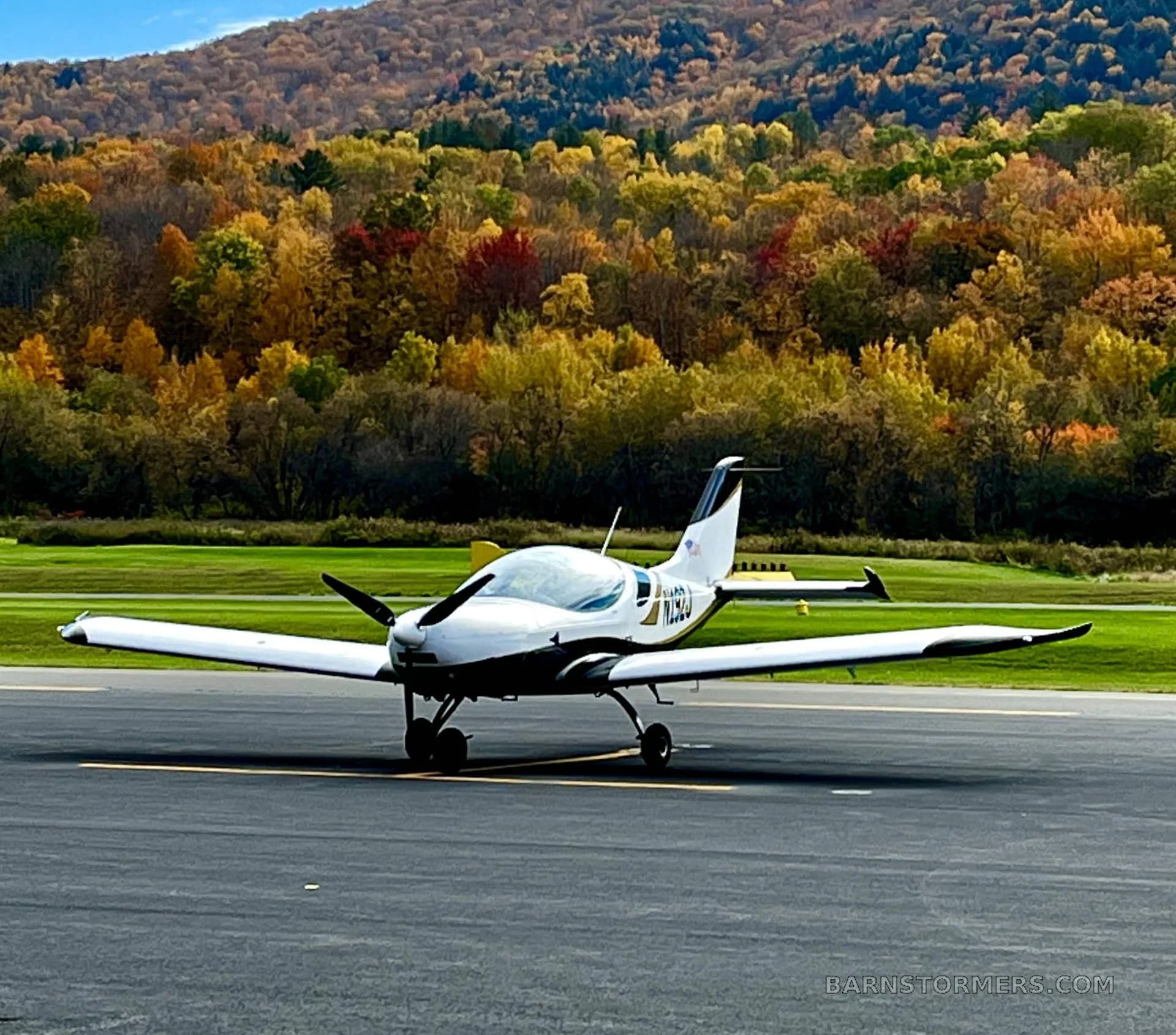A white and black aircraft with registration N192JG on a tarmac with a vibrant autumn forest in the background.