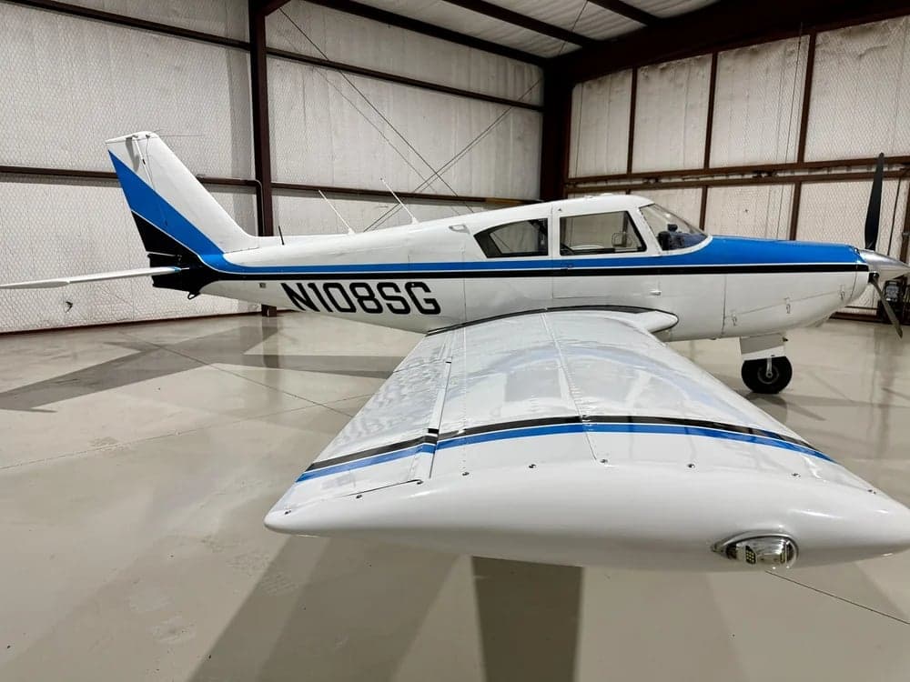 A white and blue Piper PA-28-140 Cherokee is parked in a hangar, showing the tail and wing.
