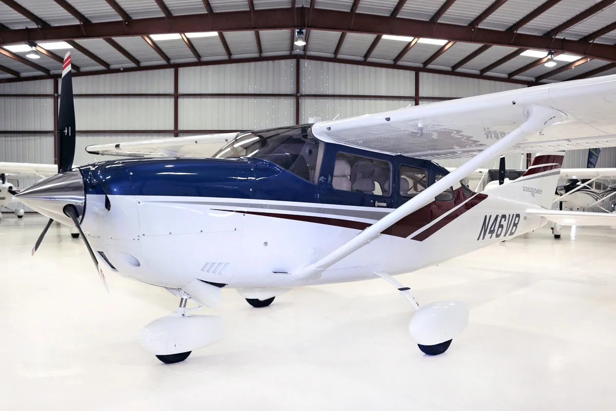 A white and blue Cessna Stationair TC aircraft (N46VB) with maroon accents is parked in a hangar, viewed from the front-left.