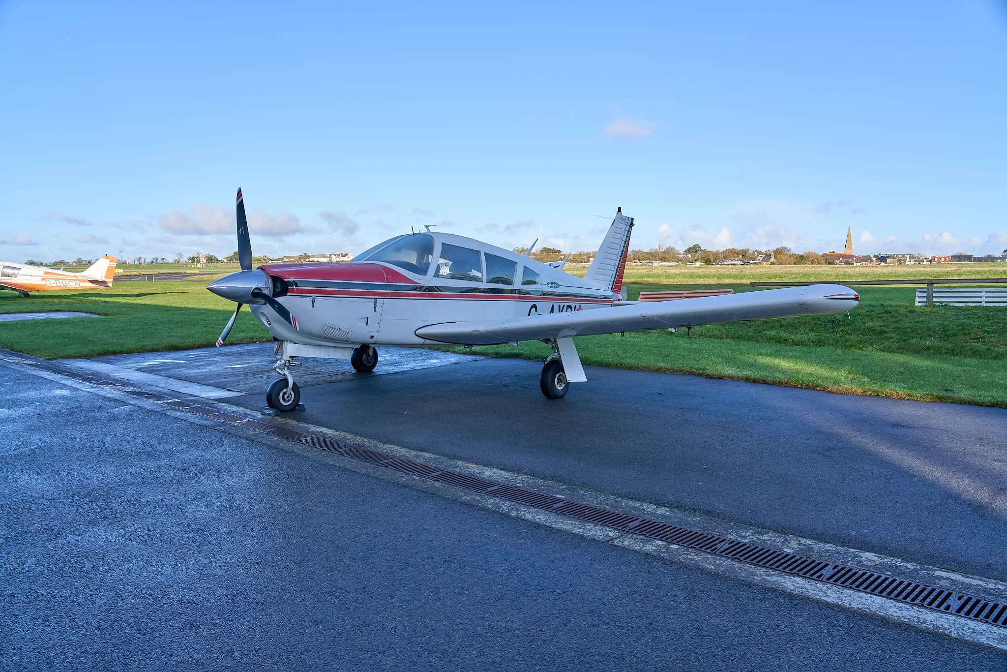A white and red single-engine propeller aircraft parked on a wet tarmac with grass and buildings in the background.