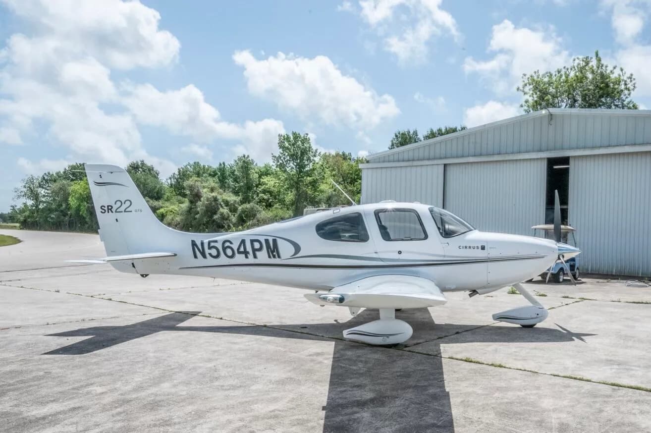 A white Cirrus SR22 GTS aircraft with N564PM registration parked on a concrete ramp next to a hangar.