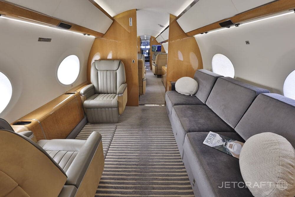 The rear section of a business jet cabin featuring a grey sofa, a single light brown leather seat, and wood paneling.