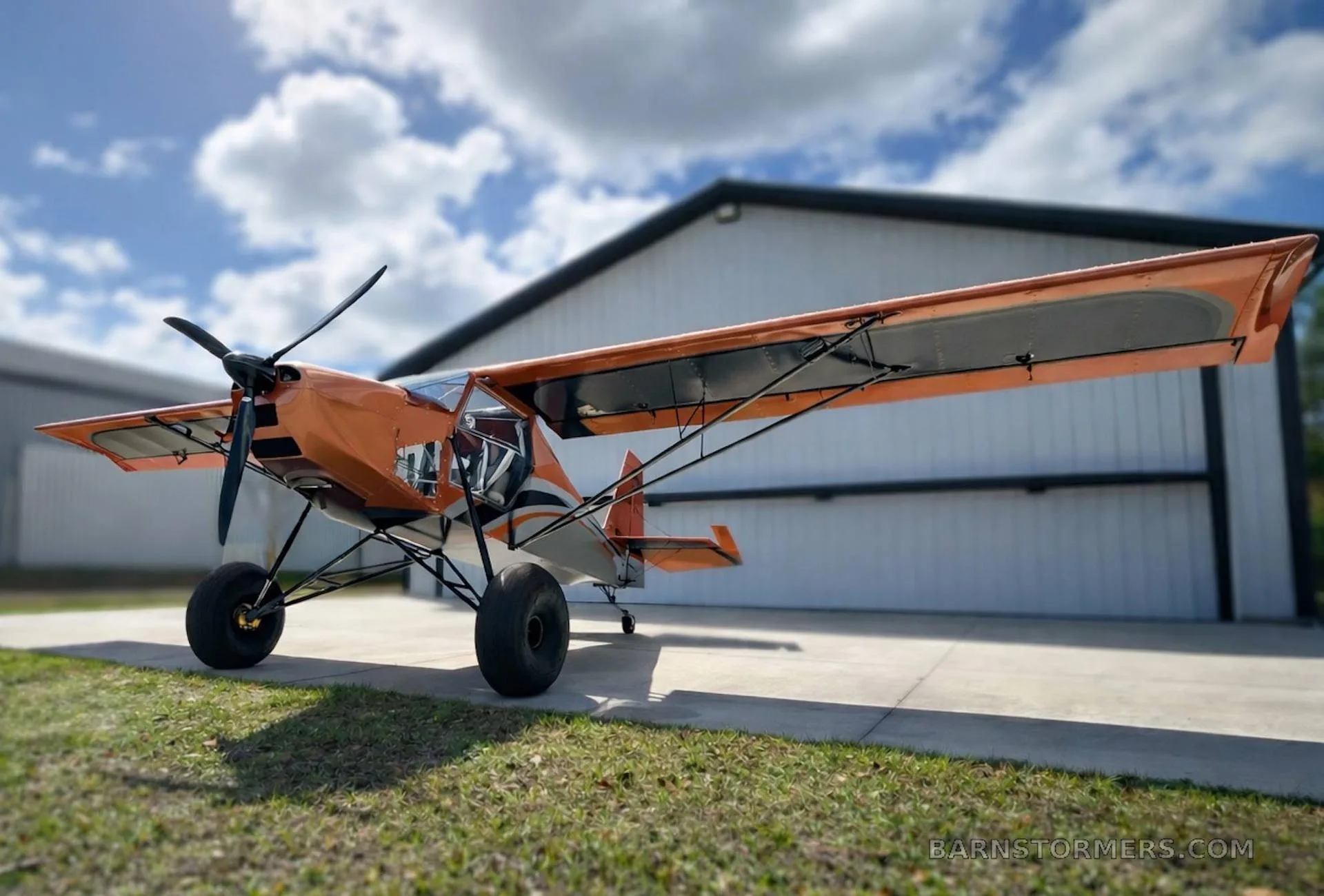 A small, orange and grey bush plane with large tires is parked on concrete next to a hangar under a partly cloudy sky.