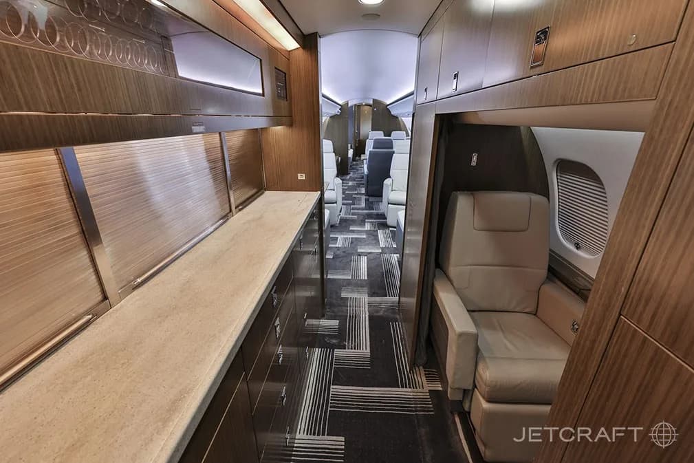 A view of the luxurious galley area and forward cabin of a Bombardier Global 6000 business jet, featuring wood cabinetry and light-colored seating.