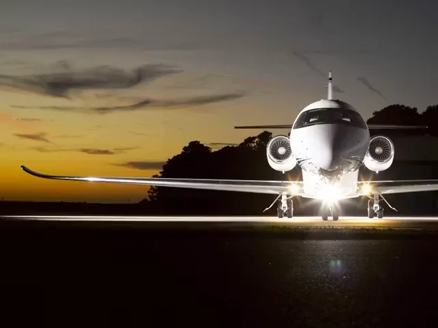 The front of a white business jet with bright landing lights on a dark tarmac at dusk.