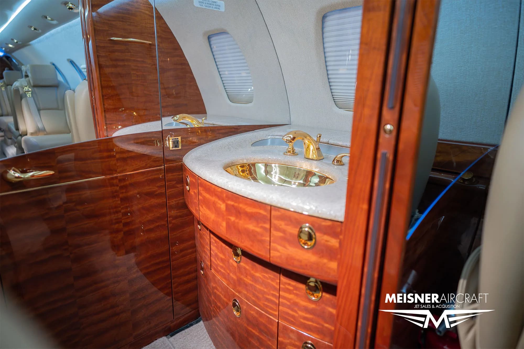 The lavatory or galley area of a business jet, featuring a sink with gold fixtures and rich wood cabinetry.