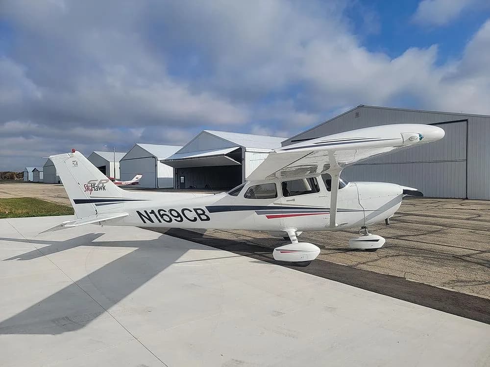 A rear-side view of the white Cessna Skyhawk N169CB with black and red stripes, parked on an airport ramp with multiple hangars in the background.