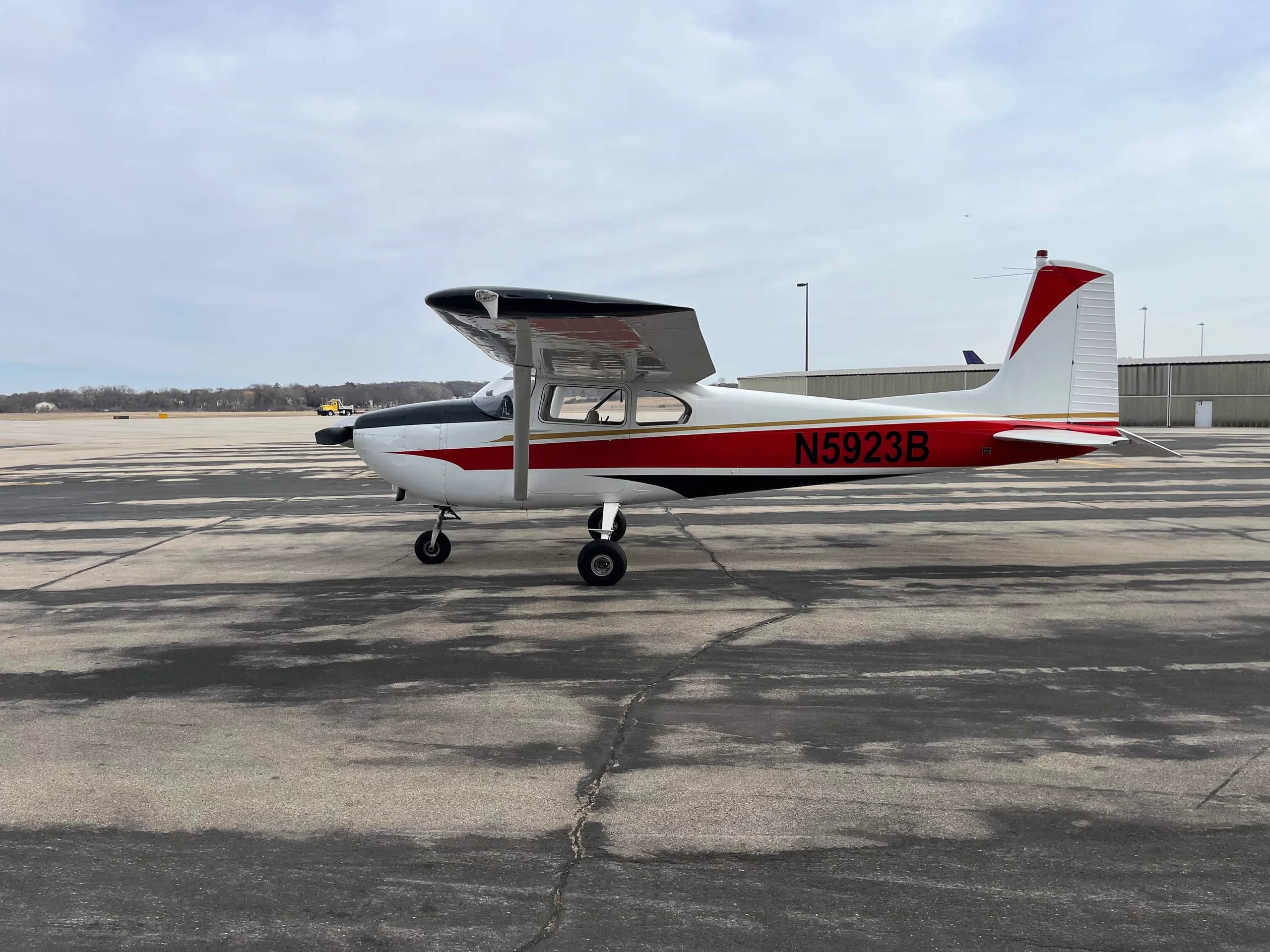 A side view of a white Cessna 182 aircraft with red and black stripes and registration N5923B, parked on a wide asphalt apron.