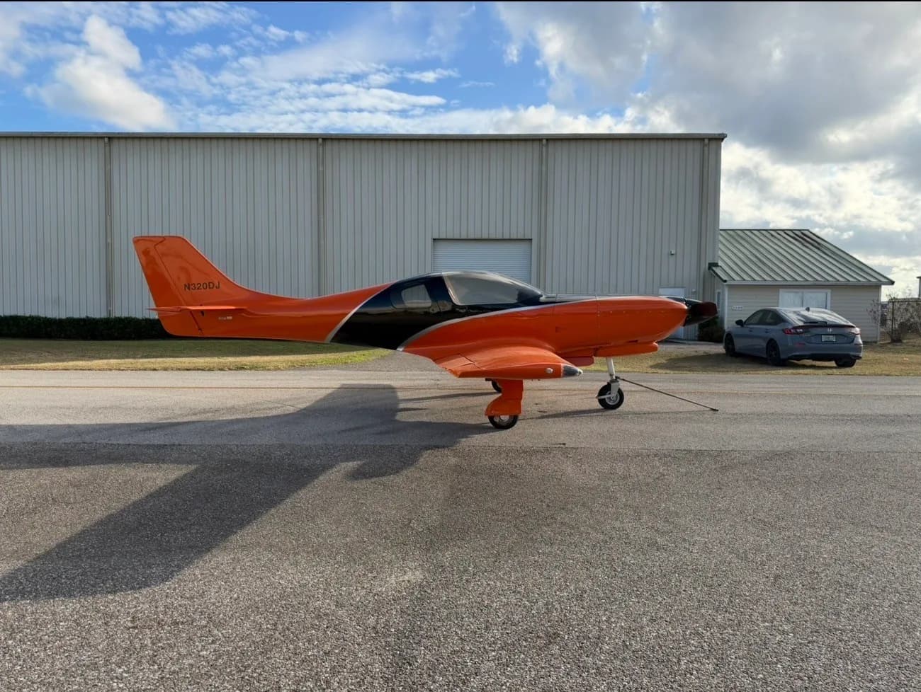 A side view of the orange and black experimental aircraft, N320DJ, parked on an airfield tarmac next to a hangar.