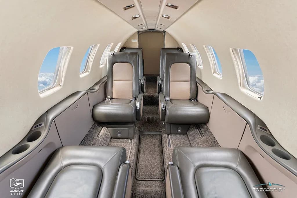 The interior cabin of a business jet features grey and tan leather seats with windows showing clouds outside.
