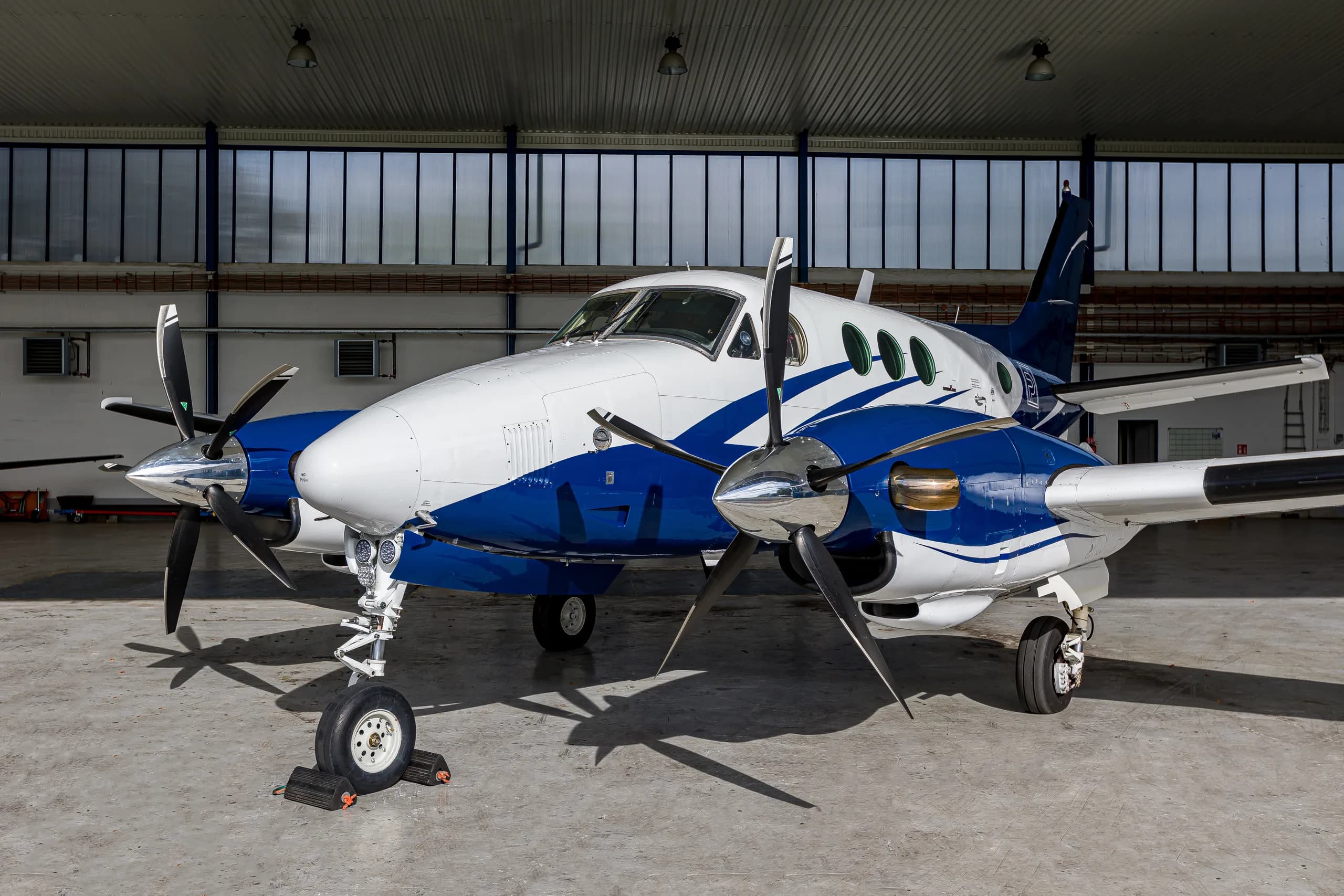 A three-quarter front view of a white and blue twin-engine turboprop aircraft, likely a King Air, parked inside a hangar.