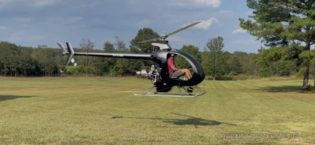 A black single-seat helicopter with a pilot hovers low over a grassy field with trees in the background.