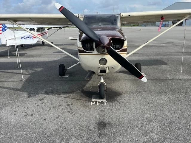 Front view of a brown and white Cessna aircraft with a propeller, parked on an asphalt tarmac.
