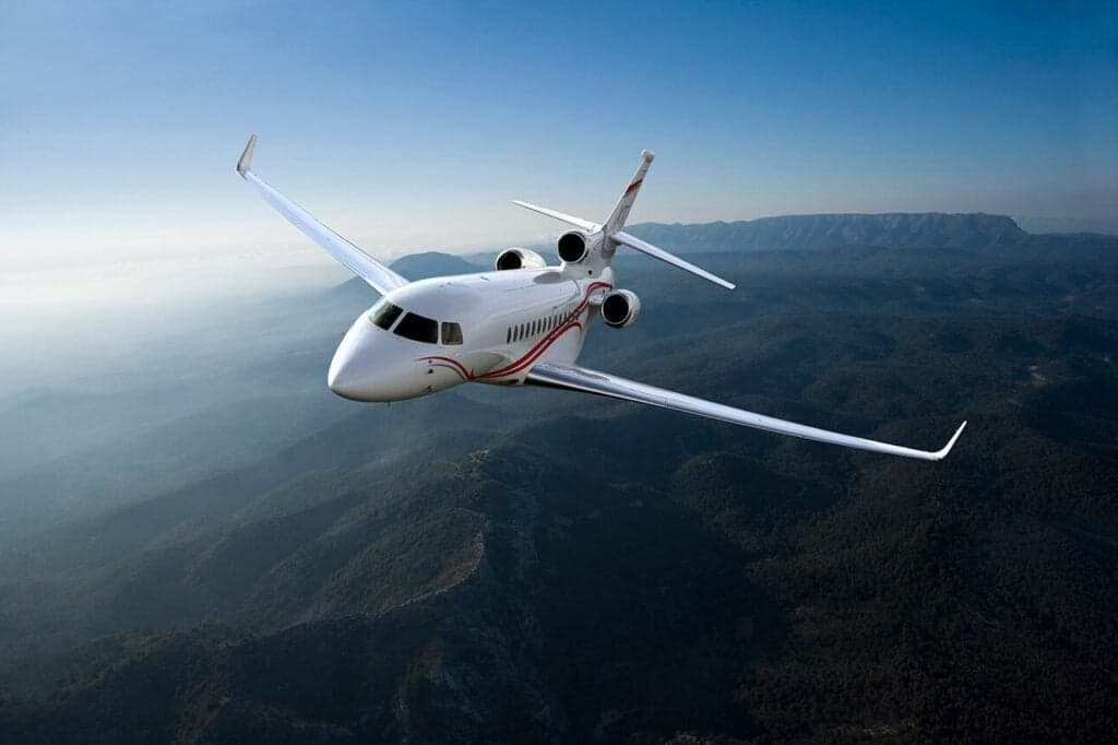 A white and red private jet flies over a mountainous landscape.