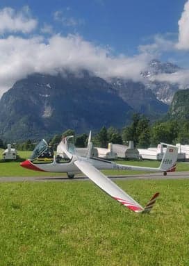 HB-2560 glider parked on a grassy field with mountains in the background.