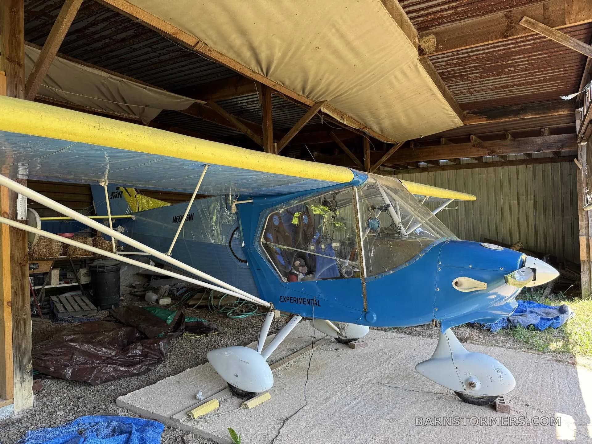 A blue and yellow experimental aircraft with white floats is parked inside a rustic shed, viewed from the front-right.