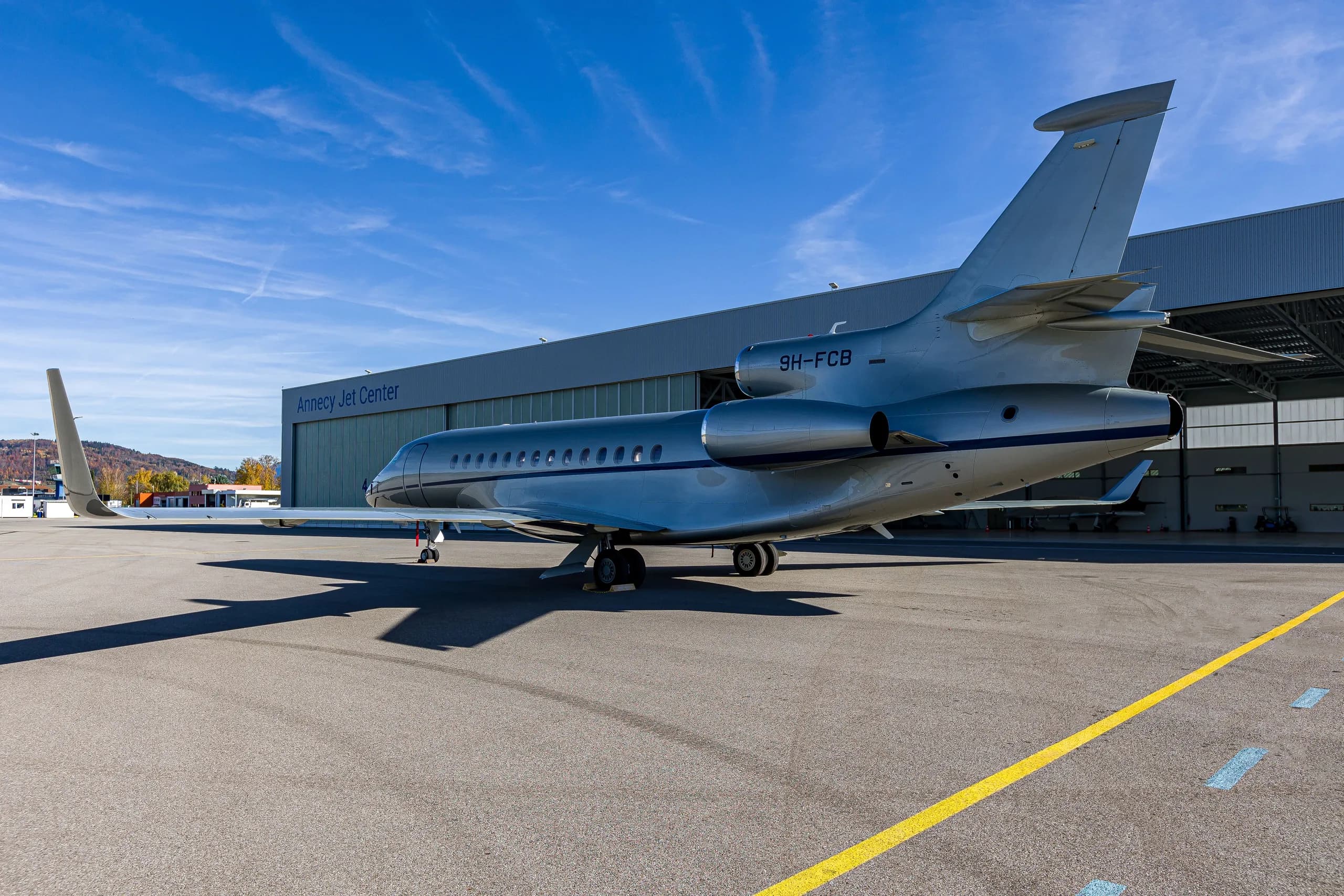 A silver Dassault Falcon 900EX business jet with blue stripes parked on an airfield in front of the Annecy Jet Center hangar.