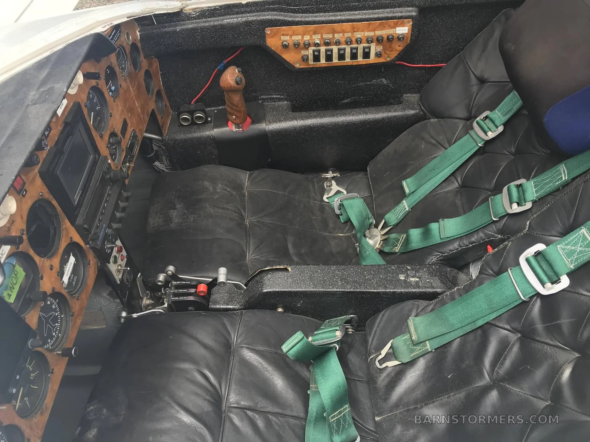 The interior of a small aircraft showing two black leather seats with green harnesses and a wooden instrument panel on the left.