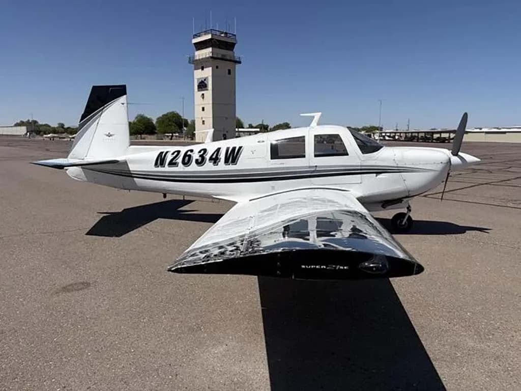 A full side view of the white Mooney M20 aircraft N2634W parked on an airfield, with an air traffic control tower in the background.