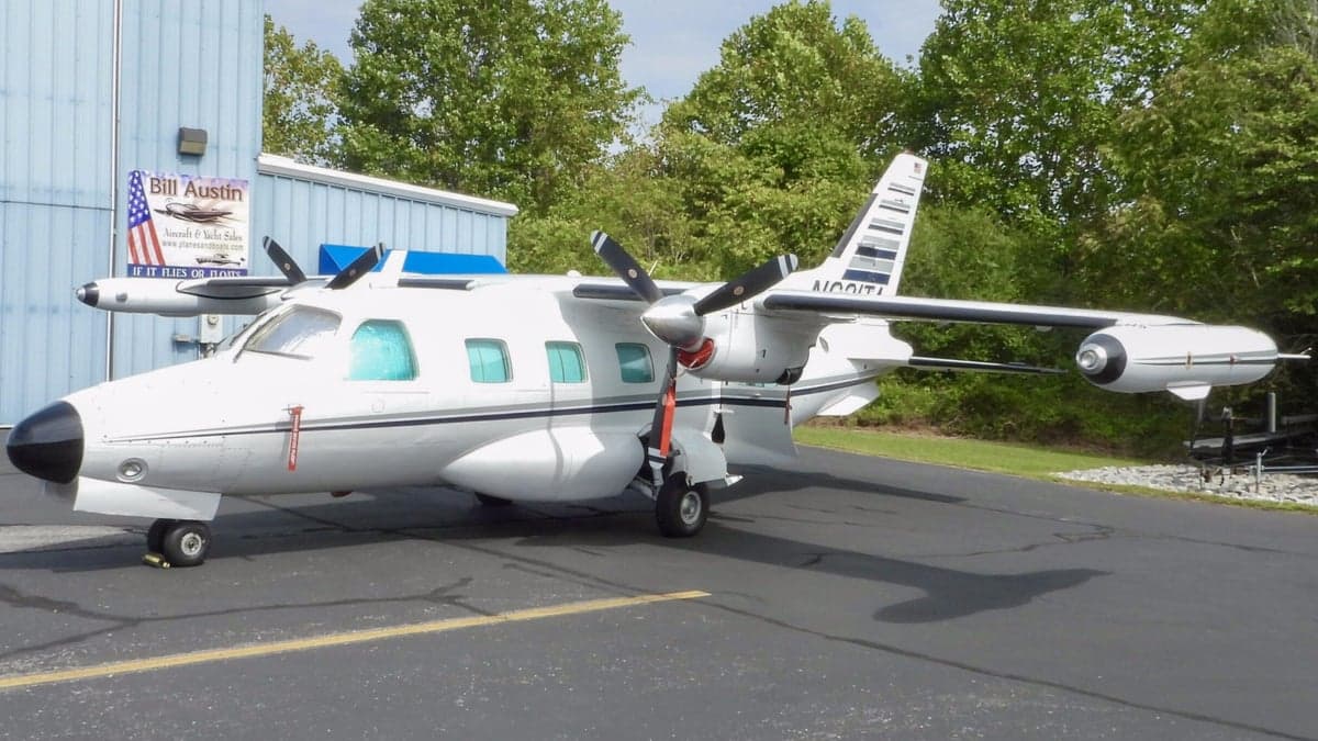 A white Mitsubishi MU-2 aircraft with black and grey stripes on the tail, parked on an asphalt ramp next to a blue building.