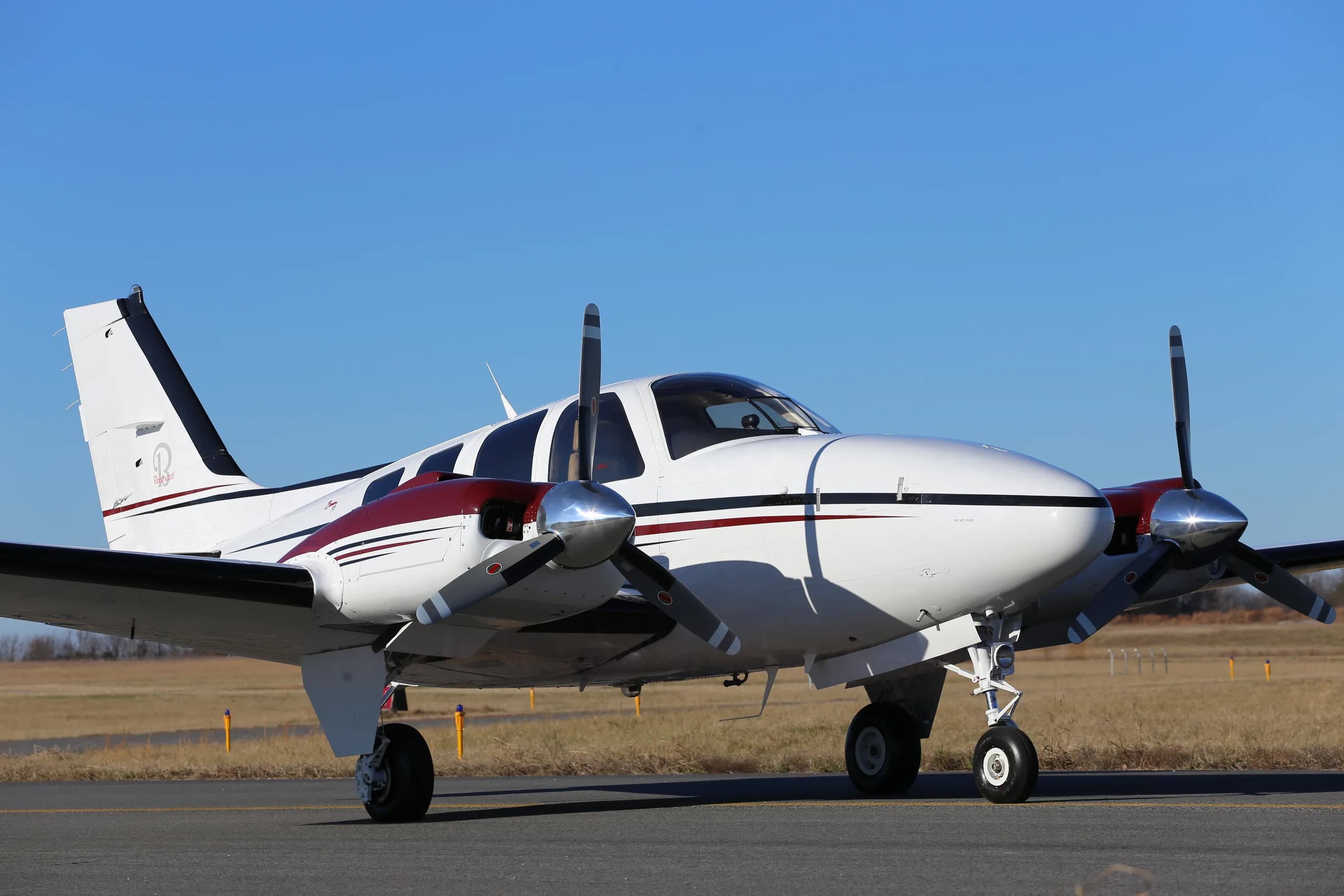 A white twin-engine propeller aircraft with maroon and black stripes sits on a tarmac under a clear blue sky.