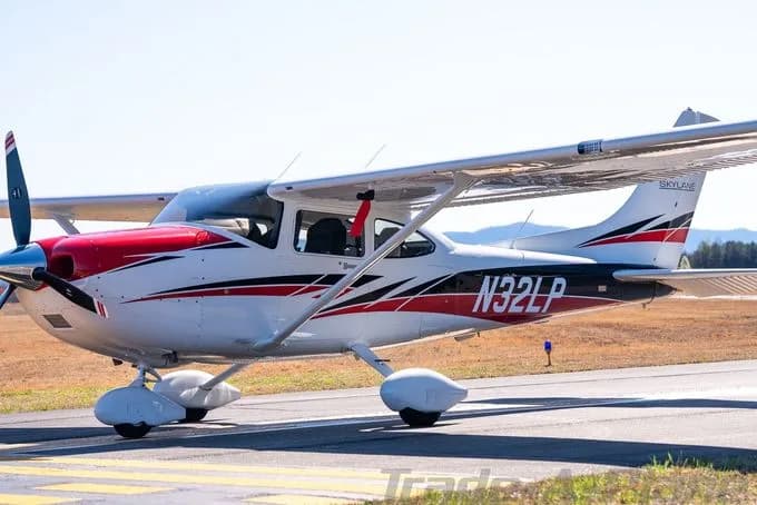 A white Cessna Skylane with red and black stripes is parked on an airfield under a clear sky.