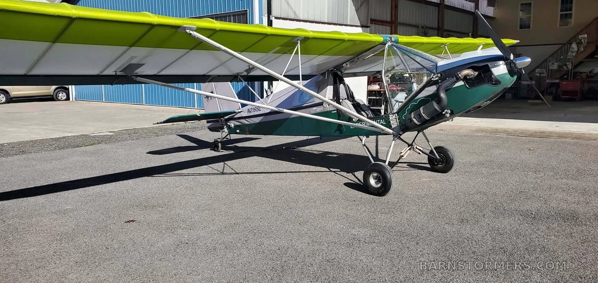 A three-quarter view of the green and white experimental aircraft, showing its high wing and tail section, parked on asphalt.
