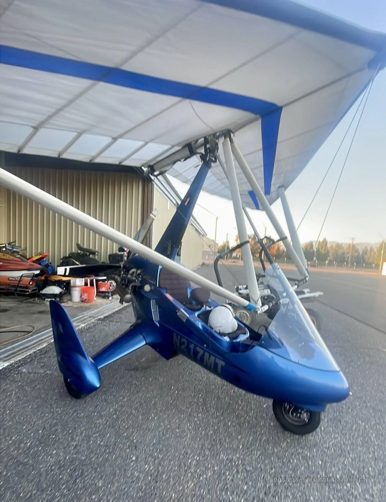 A blue ultralight aircraft with registration N217MT is parked on an airfield in front of an open hangar.