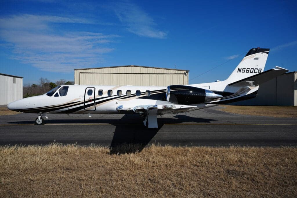 A white Cessna Citation business jet with black and gold stripes is parked on an airfield tarmac with hangars in the background.