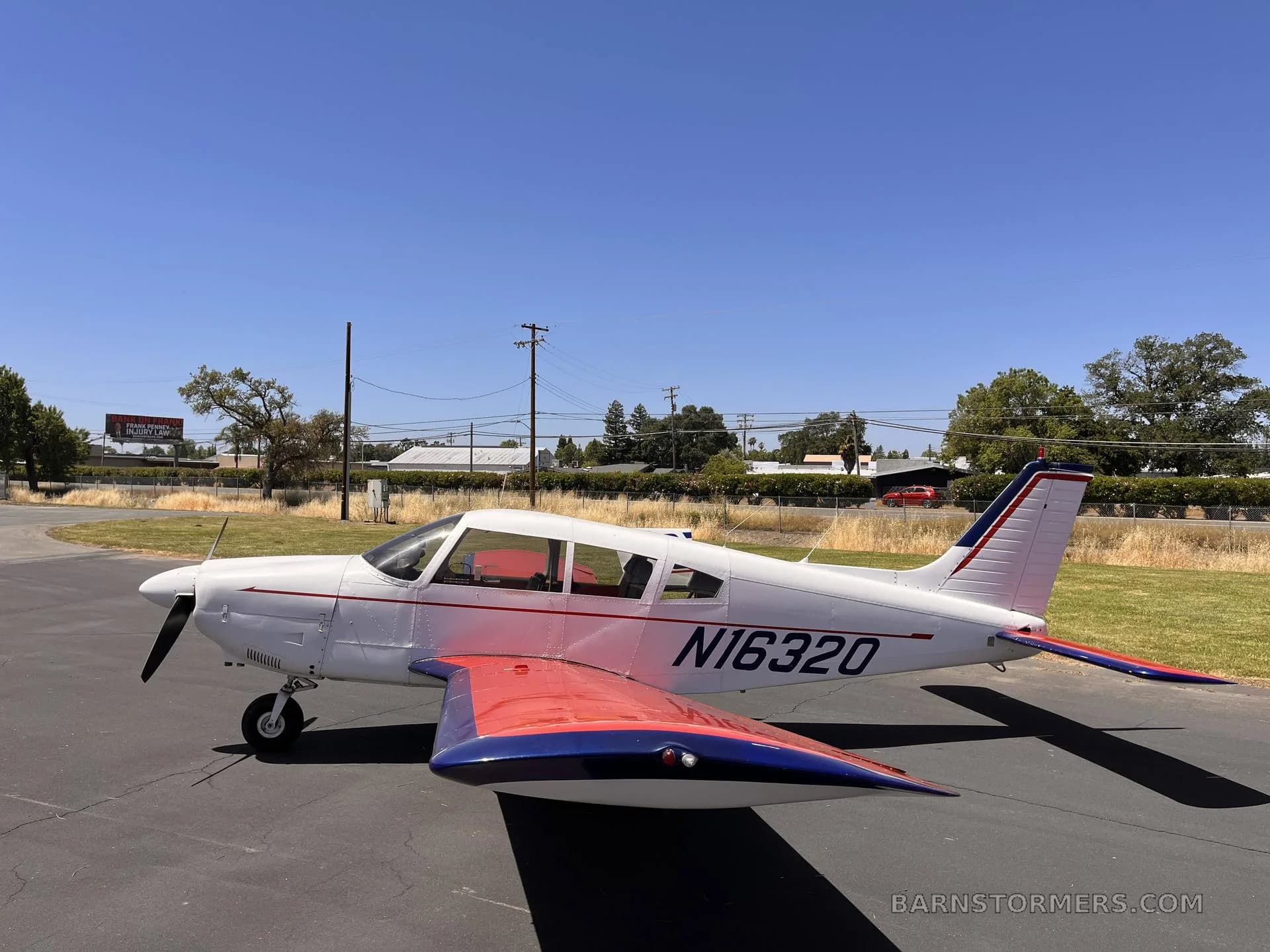 A white Piper Cherokee aircraft with red and blue accents on the wings and tail, parked on an asphalt ramp under a clear blue sky.