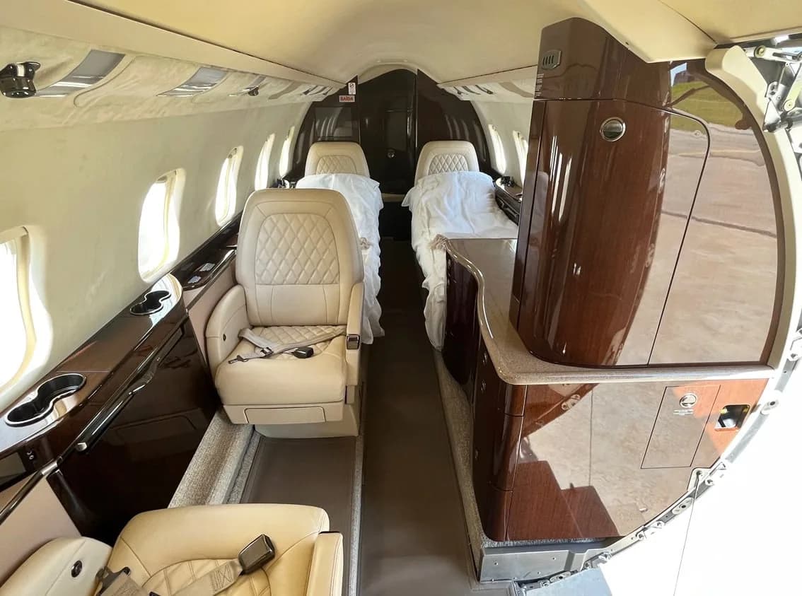 The business jet cabin shows cream leather seats, some covered in white sheets, and dark wood accents along the walls.