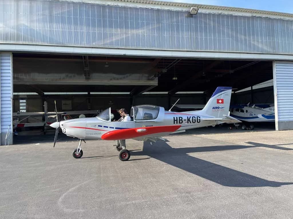 A silver Sonaca 200 aircraft with red and blue accents and registration HB-KGG is parked outside a hangar.