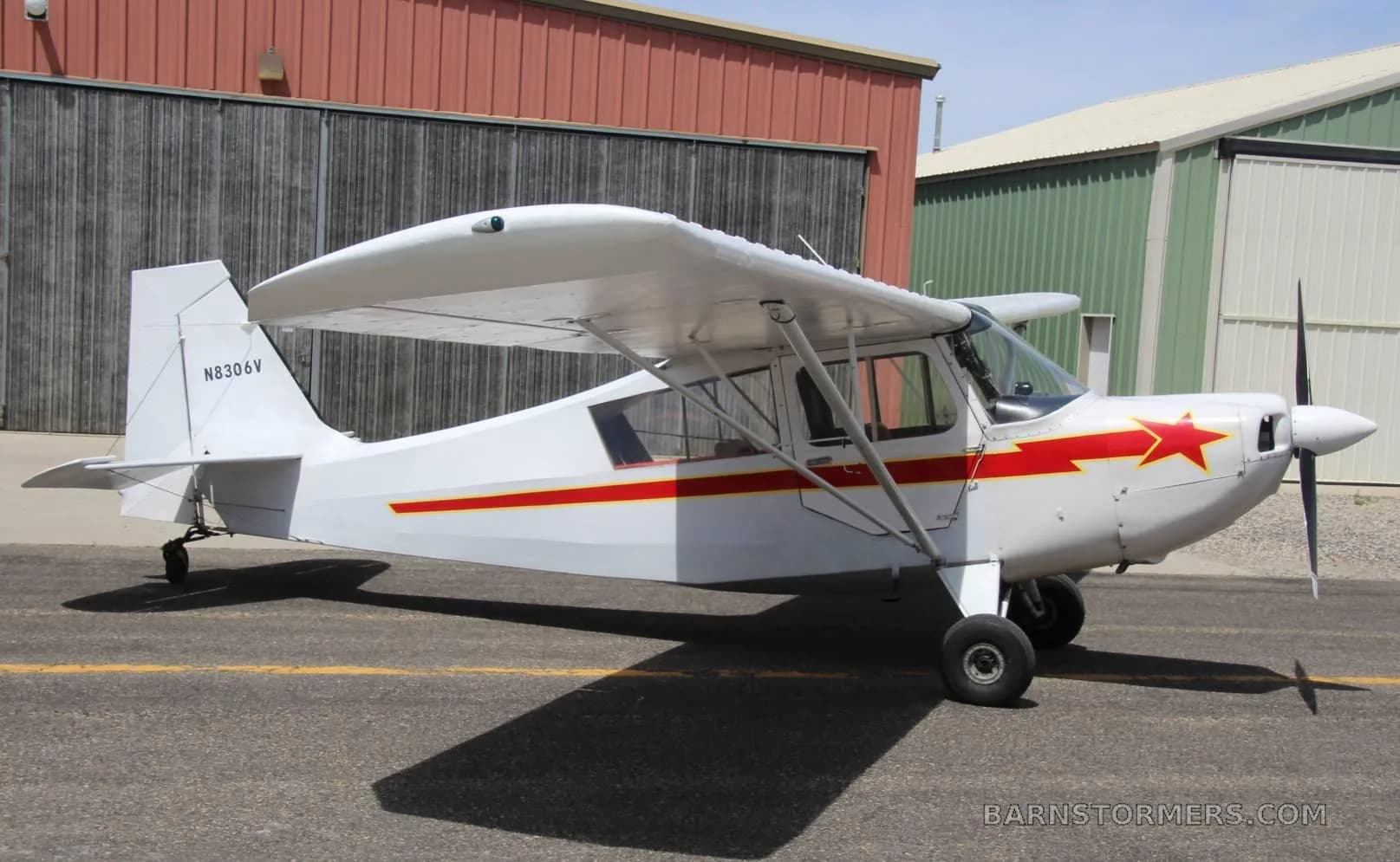 A white single-engine high-wing aircraft with red stripes and a red star on the cowl, parked on an asphalt ramp next to hangars.