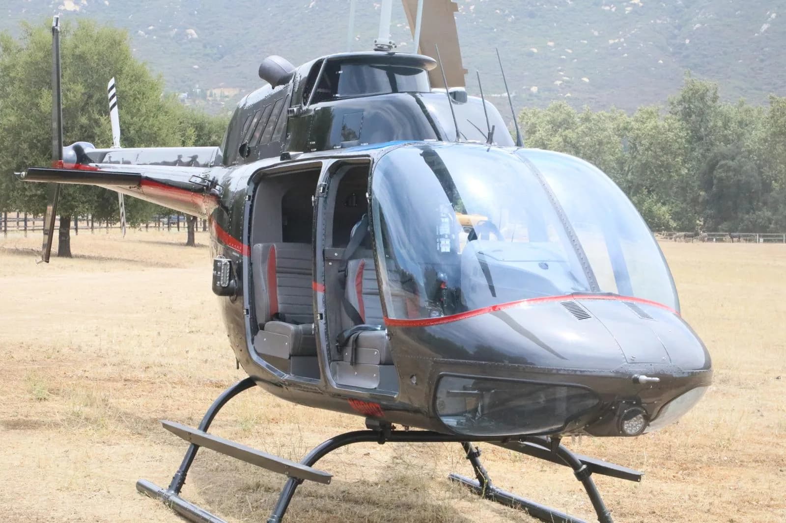 A black Bell 206 JetRanger helicopter with open doors and red accents sits in a grassy field with trees and hills.