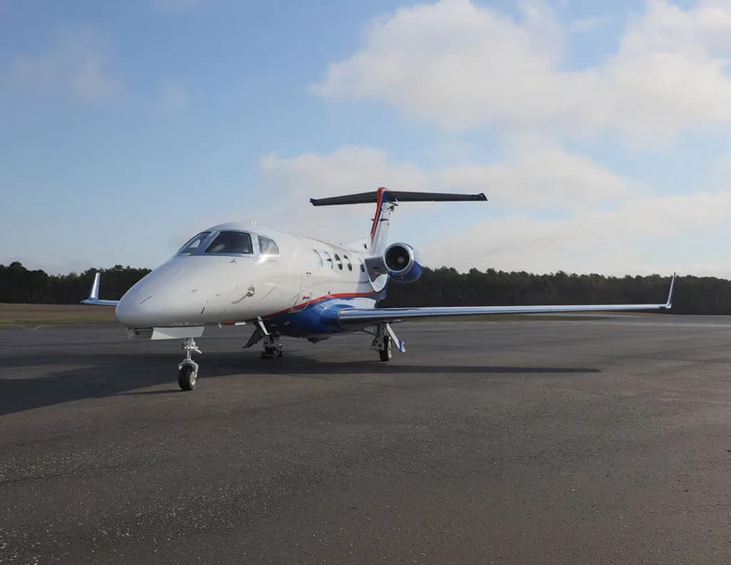 A white and blue Embraer Phenom 300 business jet is parked on an asphalt runway, viewed from the front-left.