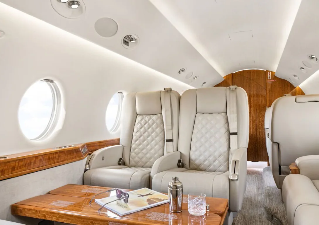 The interior of a private jet cabin shows two beige quilted seats next to a wooden table with a magazine and drink.
