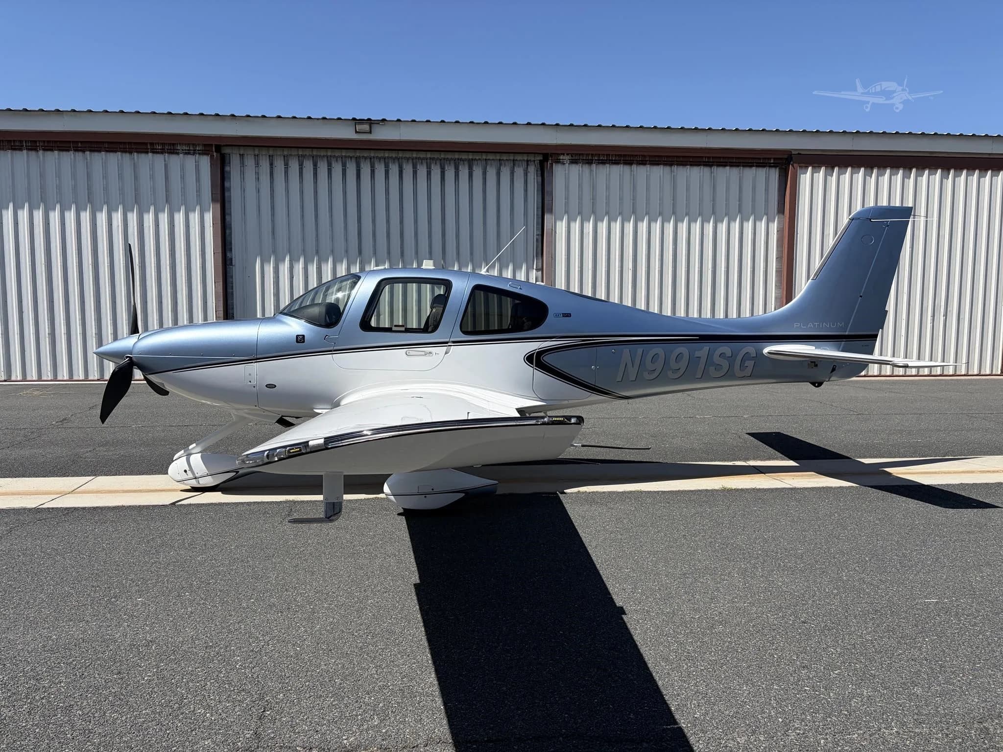 A blue and white Cirrus SR22 aircraft, registration N991SG, is parked on an asphalt tarmac in front of a hangar under a clear sky.