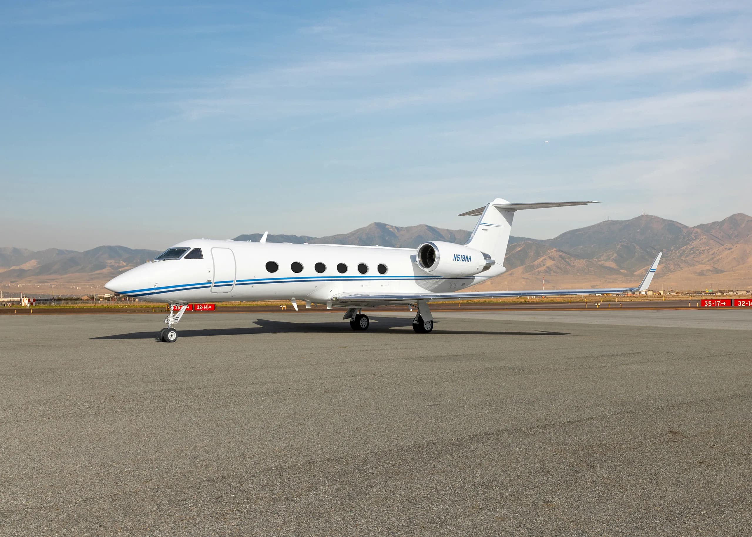 A white Gulfstream G550 business jet with a blue stripe on the tarmac, with mountains in the background.