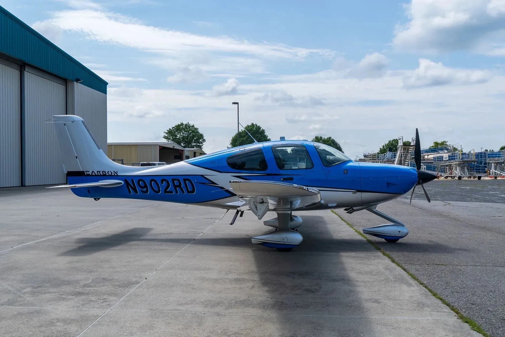 A blue and silver Cirrus SR22 aircraft with its doors closed, parked on an airfield next to a hangar.
