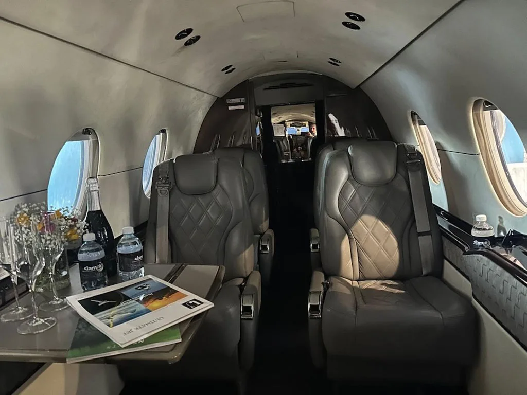 The luxurious interior cabin of a private jet features two grey leather seats, a table with drinks, and magazines.